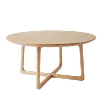 Angled view of the Astrid Round Coffee Table in natural ash, highlighting the chamfered edge of the circular top and the unique curved cross-base.