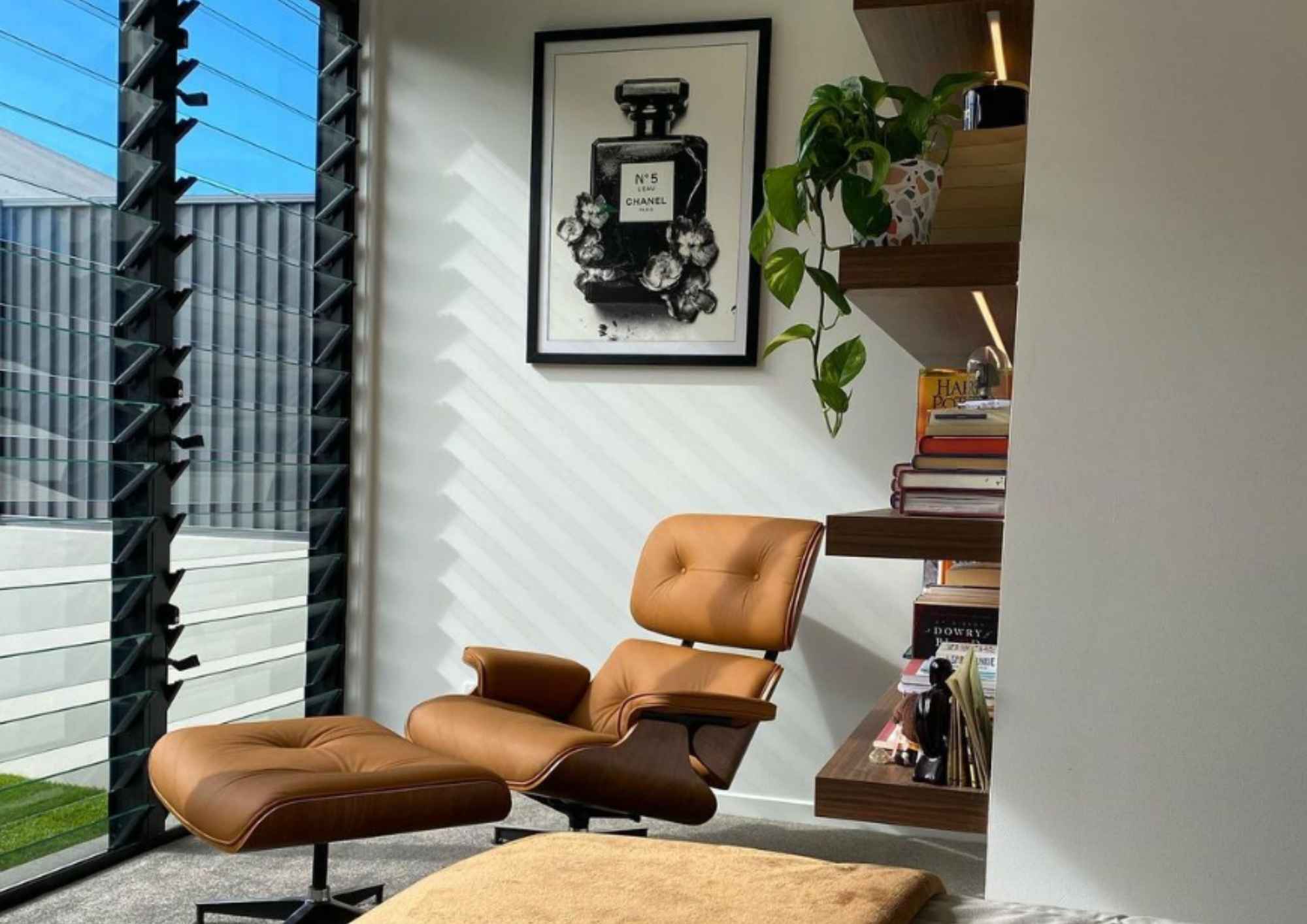 Tan leather Replica Charles Eames Lounge Chair and Ottoman in a modern living room beside a window and bookshelf.
